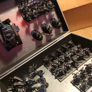 May include: A black and silver metal case containing a variety of miniature figures. The figures are arranged in rows and columns, and appear to be made of plastic or metal. The case is open, revealing the figures inside.