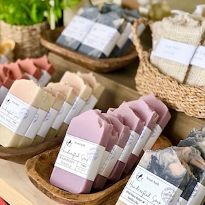 May include: Assortment of handcrafted vegan soap bars in various scents and colors, including lavender, raspberry vanilla, and charcoal. The soaps are displayed on wooden trays and baskets.