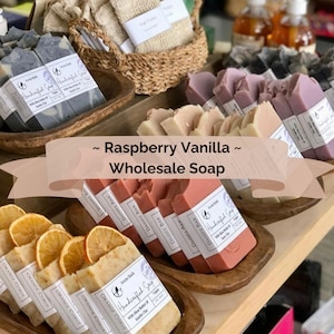 May include: Assortment of handcrafted soap bars in various scents and colors, including orange citrus, raspberry vanilla, and daisies. The soap bars are displayed in wooden bowls on a wooden table. The text "Raspberry Vanilla - Wholesale Soap" is displayed on the table.