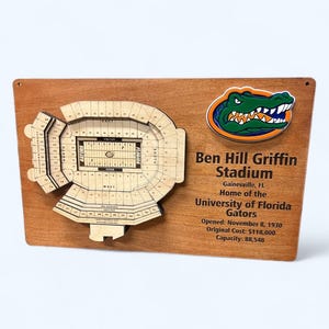 May include: A wooden sign with a 3D model of Ben Hill Griffin Stadium, home of the University of Florida Gators. The sign includes the stadium's name, location, opening date, original cost, and capacity. A green and orange alligator logo is featured in the top right corner.