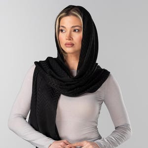 May include: A black headscarf and scarf set. The head covering is draped over the head and shoulders, with a textured pattern. The scarf is wrapped around the neck. The person is wearing a light gray long-sleeved shirt.