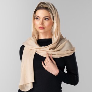 May include: A beige headscarf with a subtle pattern and delicate embellishments. The scarf is draped over the head and shoulders, creating a soft, flowing silhouette. The person is wearing a black long-sleeved top.