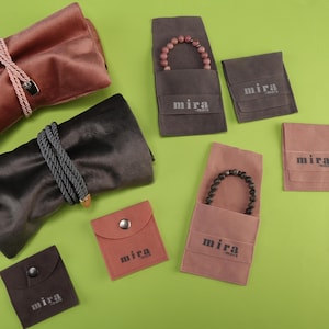 Puede incluir: Una variedad de artículos para guardar joyas en tonos marrón y gris, que incluyen estuches enrollables de terciopelo y pequeñas bolsas. Algunas bolsas contienen pulseras de cuentas. La marca "mira" está impresa.