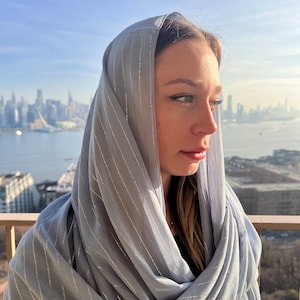May include: A woman wearing a light blue scarf with silver stripes. The scarf is draped over her head and shoulders. She is looking off to the side, and the city skyline is visible in the background.