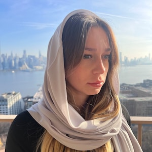 May include: A woman wearing a light gray scarf over her head and shoulders. The scarf is draped loosely around her neck and falls down her back. She is looking off to the side with a thoughtful expression.