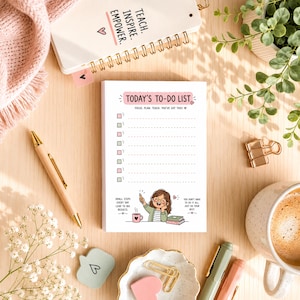 May include: A white notepad with the text "TODAY'S TO-DO LIST" and a cartoon illustration. A gold pen, a cup of coffee, and a small notebook are also visible. The notepad has a pink border and a spiral-bound notebook is in the background.