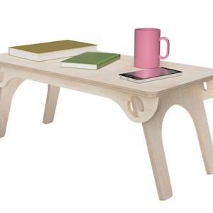 Puede incluir: Un escritorio portátil de madera clara con patas curvas. En el escritorio hay dos libros, una taza rosa y un teléfono inteligente. El escritorio está diseñado para usar en una cama o sofá.