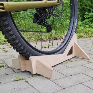 May include: A wooden bicycle stand supporting a mountain bike's rear wheel. The stand is made of light-colored wood and has a geometric design. The bike tire is black with a SCHWALBE label. The background is a paved surface and greenery.