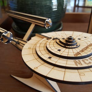 Puede incluir: Un modelo de madera de la nave espacial USS Enterprise de Star Trek. El modelo está pintado en un color madera claro y tiene detalles negros. El nombre de la nave, "USS Enterprise", y el número de registro, "NCC-1701", están impresos en la sección del platillo.