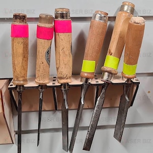 May include: A set of six wood carving chisels with wooden handles. The handles are painted with pink and yellow tape. The chisels are mounted on a wooden rack.