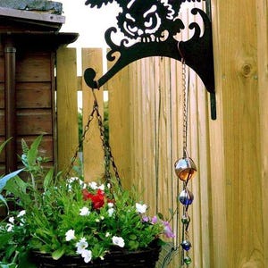 Puede incluir: Soporte de cesta colgante de metal negro con forma de búho con una cesta colgante llena de flores blancas y rojas. Una campanilla de viento decorativa cuelga debajo del soporte. El soporte está montado en una valla de madera.