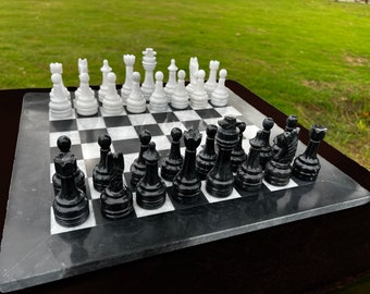 Può includere: Un set di scacchi in marmo con pezzi bianchi e neri disposti su una scacchiera. I pezzi degli scacchi sono bianchi e neri, e la scacchiera è grigio scuro con quadrati bianchi. Il set è esposto su una superficie scura con uno sfondo verde.