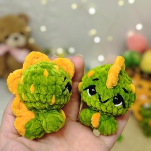 May include: Two small, plush, crocheted dinosaur figures made from soft green and orange yarn. Each creature features a rounded body, a textured surface, and orange spikes along the back. The figures have large, dark, embroidered eyes and a stitched mouth.