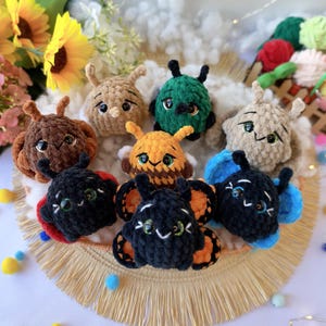 Può includere: Una collezione di otto piccole creature di peluche all'uncinetto, rotonde, con antenne e grandi occhi espressivi. I peluche presentano vari colori tra cui marrone, verde, beige, arancione, nero e blu, alcuni con motivi simili ad ali.