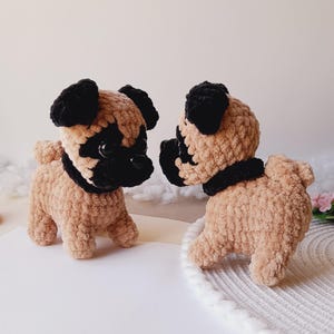 May include: Two small, handmade crochet pug dog figurines made from soft, tan-colored yarn with black yarn accents on the ears, face, and around the neck. The pugs have dark, round eyes and are positioned facing each other on a white surface.