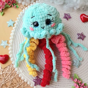 Pode incluir: Um brinquedo de água-viva de crochê com um corpo azul claro, tentáculos amarelos, vermelhos e rosados. A água-viva tem um rosto sorridente com olhos pretos e bochechas rosadas.