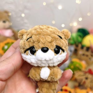 May include: A small, handmade crochet plush toy shaped like an otter, crafted from soft, light brown and white yarn. The figure features large, glossy black safety eyes, a small black nose, and a white muzzle, with a textured, fuzzy appearance.