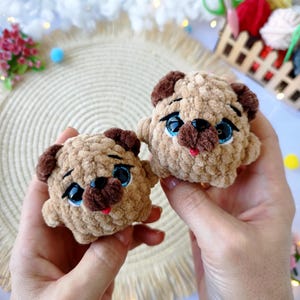 May include: Two handmade, plush pug dog toys. The toys are light brown with darker brown accents on the ears and snout. They have blue eyes and small red tongues. The toys are held in a person's hands.