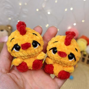 May include: Two small, round, yellow crocheted plush chickens with a soft, fuzzy texture. Each chicken features a bright red comb on top, a red beak, and large, glossy black eyes with white highlights. They have small red feet and wings.