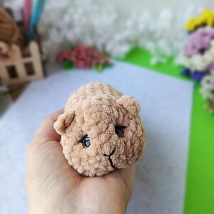 May include: A small, handmade, light brown crocheted animal toy with black eyes and a smiling mouth. The plush toy is held in a hand, showcasing its soft, textured appearance. The toy is likely a hamster or similar small animal.