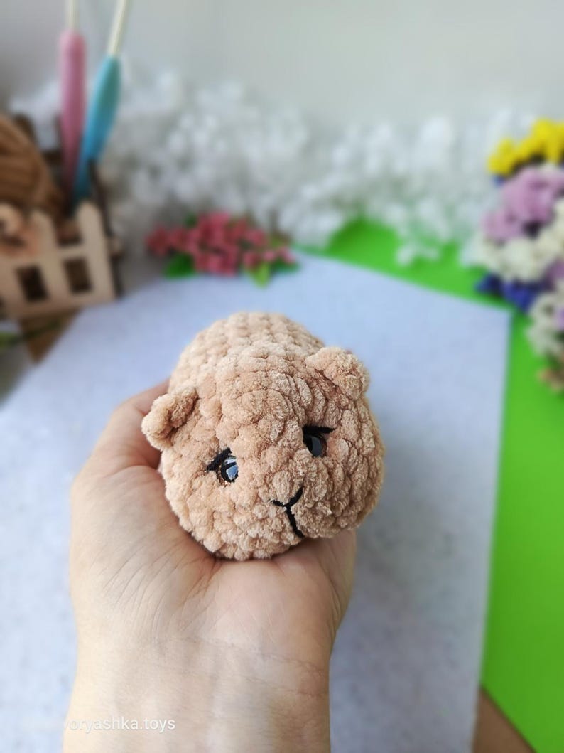 May include: A small, handmade, light brown crocheted animal toy with black eyes and a smiling mouth. The plush toy is held in a hand, showcasing its soft, textured appearance. The toy is likely a hamster or similar small animal.