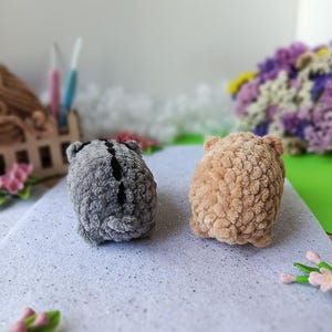 May include: Two handmade crochet animals. One is gray with a black stripe, the other is tan. Both have small ears and a rounded shape. They are placed on a white, glittery surface.