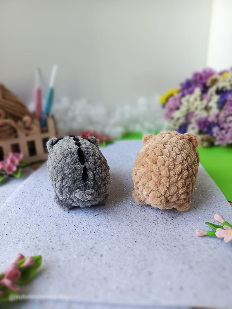 May include: Two handmade crochet animals. One is gray with a black stripe, the other is tan. Both have small ears and a rounded shape. They are placed on a white, glittery surface.