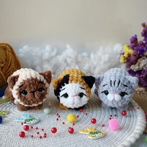 Può includere: Tre figurine di gatto all'uncinetto fatte a mano in marrone e crema, tartarugato e grigio. Ogni gatto ha tratti del viso dettagliati ed è seduto su una superficie bianca e strutturata. Elementi decorativi: perline rosse e piccoli ritagli di legno.