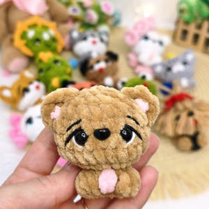 May include: A small, tan, crocheted bear figurine with a soft, plush texture. The bear features large, glossy black eyes with embroidered black eyelashes, small pink inner ears, and a small pink patch on its chest, with a black plastic nose.