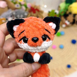 May include: A small, handmade crochet fox plush toy featuring soft orange yarn, a white muzzle and chest patch, black ears with white accents, and black paws. The fox has large, glossy black safety eyes and a small black nose, with a textured finish.