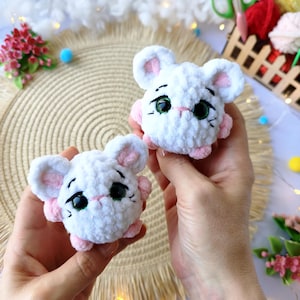 May include: Two handmade white crochet mice with pink accents. Each mouse has large black eyes, pink ears, and pink paws. The mice are made of soft, textured yarn and are designed to look like cartoon characters. The background includes craft supplies.