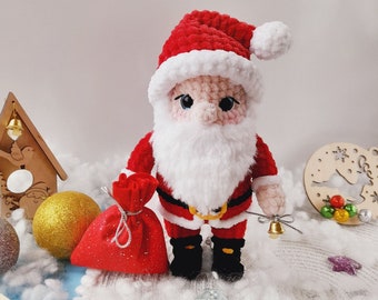 Pode incluir: Um boneco de Papai Noel de crochê vestindo um chapéu vermelho com acabamento branco, um terno vermelho com cinto branco e barba branca. O boneco está segurando uma bolsa vermelha e um sino dourado.