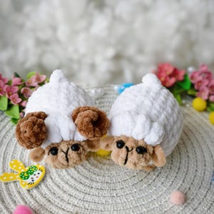 May include: Two handmade crochet sheep toys. The sheep have white bodies, brown ears, and tan faces with black eyes. They are placed on a textured surface with decorative Easter-themed accents, including yellow bunny cutouts and colourful pom-poms.