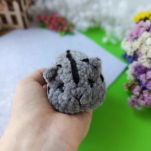 May include: A small, gray crocheted cat toy with black stripes and blue eyes. The handmade plush animal has a rounded shape and is held in a hand. The toy is a soft, cuddly item.