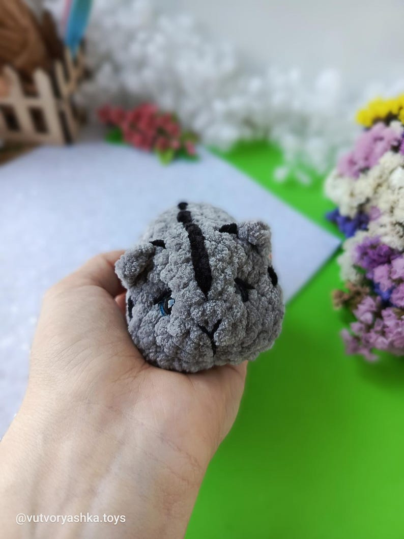 May include: A small, gray crocheted cat toy with black stripes and blue eyes. The handmade plush animal has a rounded shape and is held in a hand. The toy is a soft, cuddly item.