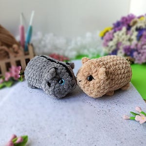 May include: Two handmade crochet hamster toys. One is gray, the other is tan. Both have small black eyes and are positioned side-by-side. The toys are made of a soft, textured yarn. The background includes flowers and a small wooden structure.