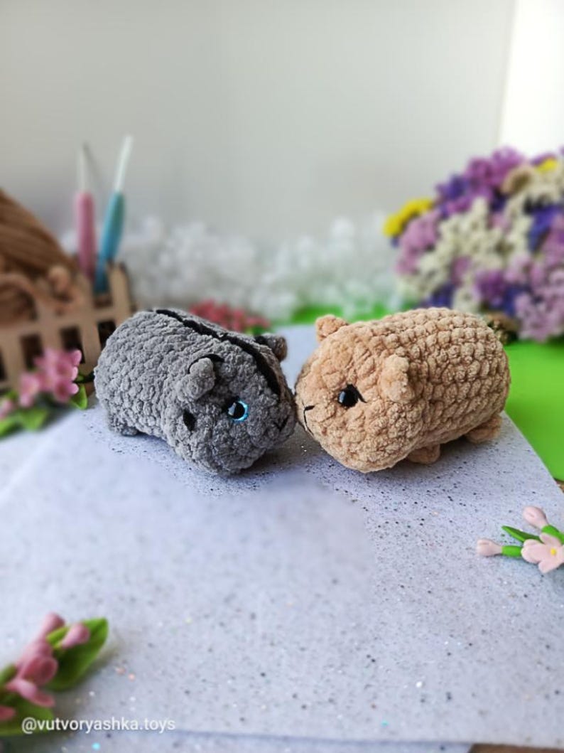 May include: Two handmade crochet hamster toys. One is gray, the other is tan. Both have small black eyes and are positioned side-by-side. The toys are made of a soft, textured yarn. The background includes flowers and a small wooden structure.