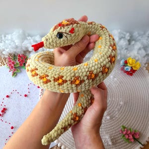 May include: A crocheted snake toy with a light brown body and orange and yellow accents. The snake has a red tongue and black eyes.