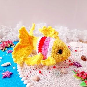 Puede incluir: Un juguete de peluche tejido a crochet con forma de pez amarillo y rosa con una franja blanca. El pez está sentado sobre una superficie de crochet blanco con cuentas rojas y conchas.
