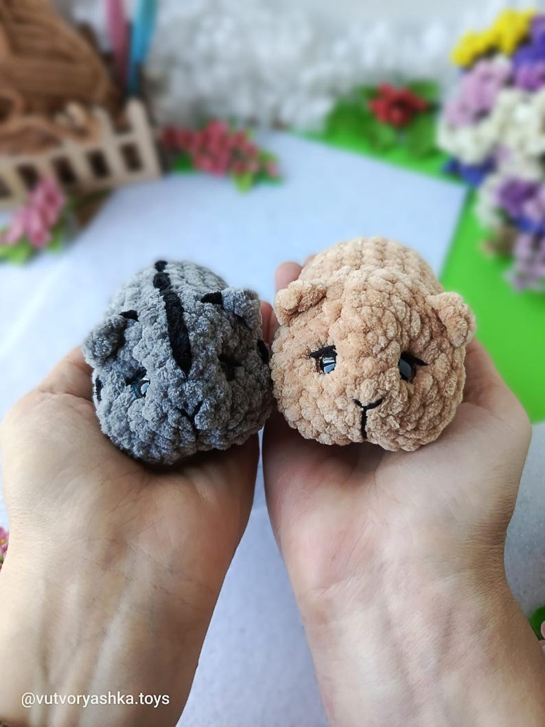 May include: Two handmade crochet animal toys. One is gray with black stripes, the other is tan. Both have small black eyes and are held in open hands. The toys are made of soft, textured yarn. The background includes flowers and a white surface.