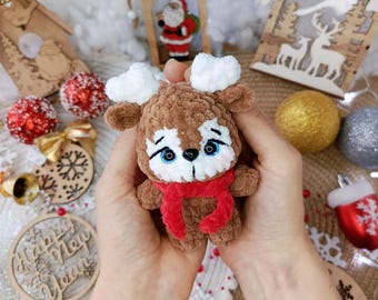 May include: A handmade brown plush reindeer with white accents, blue eyes, and a red scarf. The reindeer is held in hands, surrounded by Christmas ornaments and wooden decorations, including a Santa Claus figure.