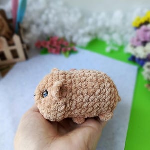 May include: A handmade, plush, light brown hamster toy with small ears and black eyes. The soft, textured toy is held in a hand, showcasing its small size and detailed craftsmanship. The background includes craft supplies and flowers.