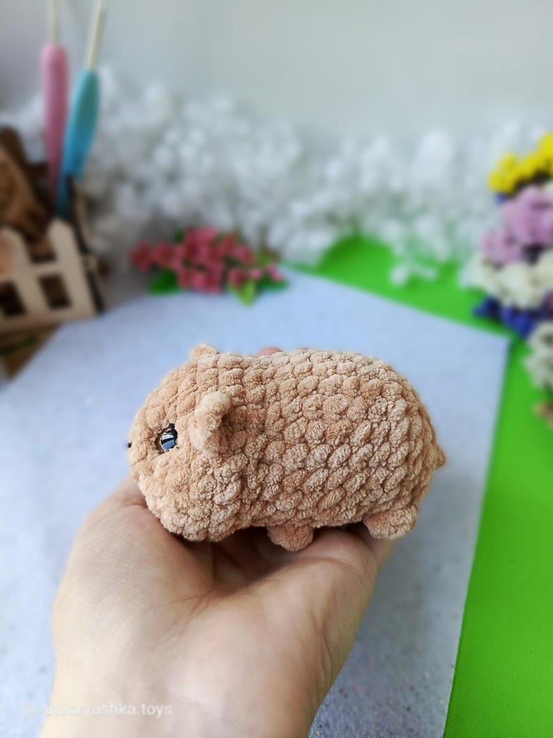 May include: A handmade, plush, light brown hamster toy with small ears and black eyes. The soft, textured toy is held in a hand, showcasing its small size and detailed craftsmanship. The background includes craft supplies and flowers.