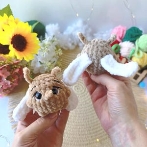 May include: Two small, round, tan-coloured crochet plush toys with white wings and antennae. The plush toys have a soft, velvety texture, dark plastic eyes, and a small protruding nose. Each toy features a rounded body and two white wings.