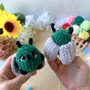 Puede incluir: Dos juguetes de peluche hechos a ganchillo con forma de escarabajos. Cada escarabajo tiene un cuerpo redondo de textura verde oscuro, dos pequeñas antenas negras y alas de color gris claro. El escarabajo de la izquierda tiene ojos grandes.