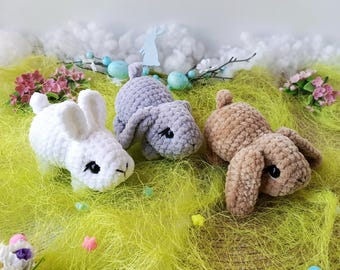 May include: Three crocheted bunny rabbits in different colors: white, gray, and brown. The bunnies are sitting on a bed of green grass.