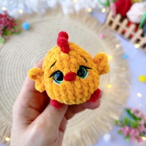 May include: A handmade, yellow crocheted chick with a red comb, beak, and feet. The chick has large, expressive green eyes with black outlines. The plush toy is round and soft, perfect for a child's toy or nursery decor.