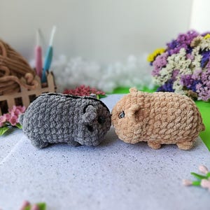 May include: Two handmade crochet guinea pig toys. One is gray, the other is tan. Both have small ears and black eyes. The toys are made with a soft, textured yarn and are sitting on a white surface.