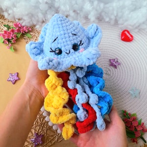 Pode incluir: Uma nuvem azul de crochê com um rosto feliz e tentáculos de fio arco-íris. A nuvem é segurada em uma mão com um coração rosa que diz "Happy" no fundo.