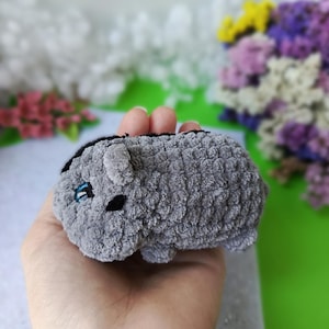 May include: A small, gray plush toy in the shape of a hamster. The toy has black eyes and a black stripe down its back. The hamster is held in a hand, with a blurred background of flowers.